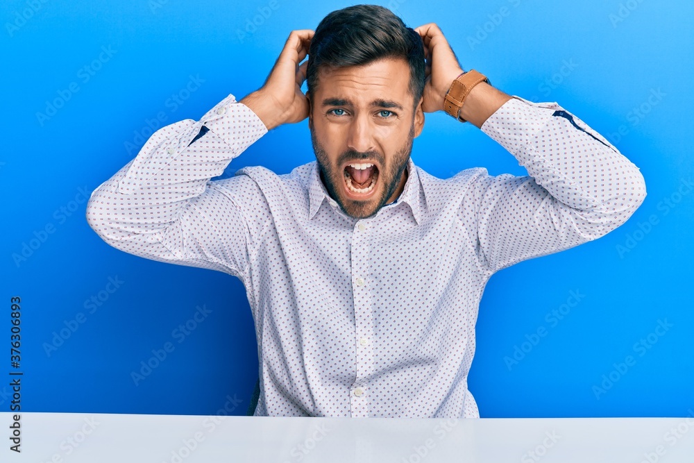 Handsome hispanic man wearing business clothes sitting on the table crazy and scared with hands on head, afraid and surprised of shock with open mouth Handsome hispanic man wearing business clothes sitting on the table crazy and scared with hands on head, afraid and surprised of shock with open mouth