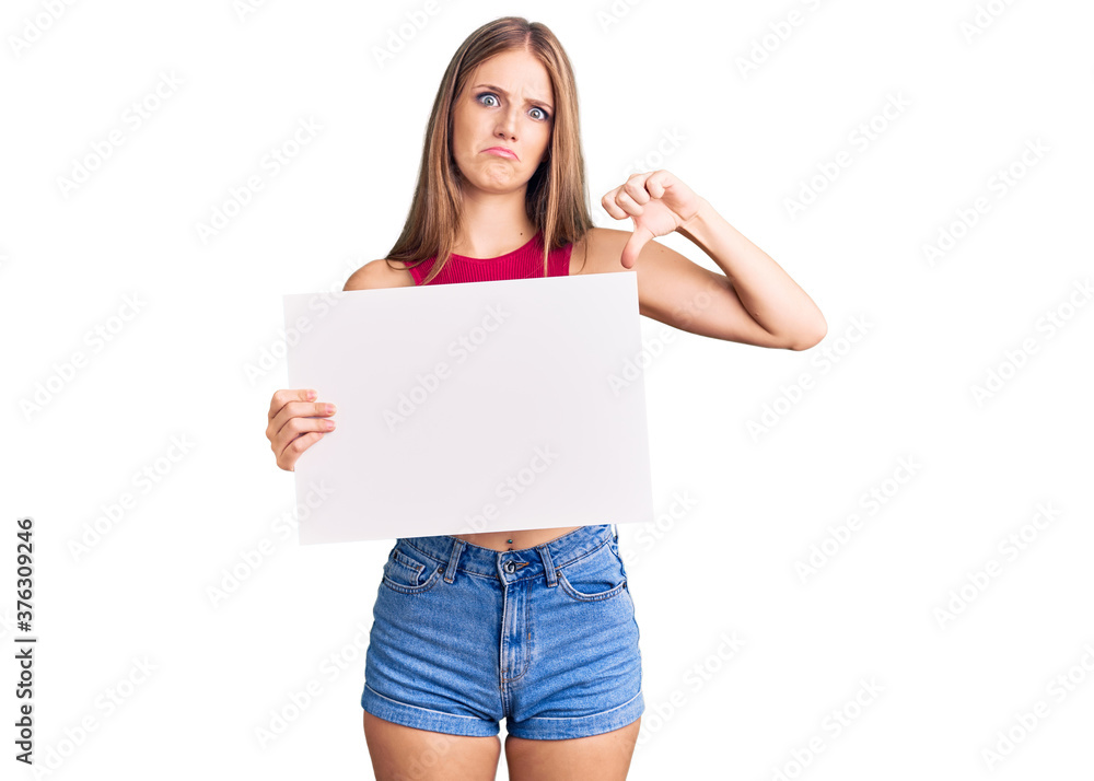 Young beautiful blonde woman holding blank empty banner with angry face, negative sign showing dislike with thumbs down, rejection concept Young beautiful blonde woman holding blank empty banner with angry face, negative sign showing dislike with thumbs down, rejection concept