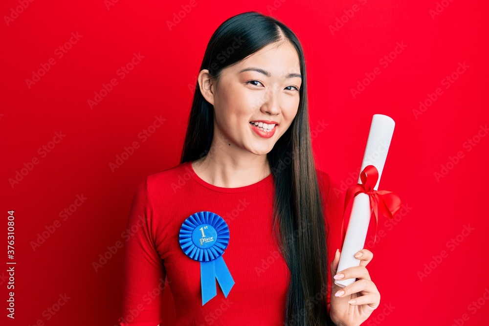 Young chinese woman wearing first place badge holding diploma looking positive and happy standing and smiling with a confident smile showing teeth Young chinese woman wearing first place badge holding diploma looking positive and happy standing and smiling with a confident smile showing teeth