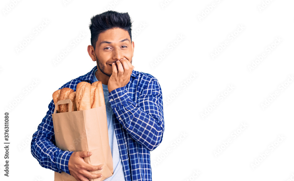 Handsome latin american young man holding paper bag with bread laughing and embarrassed giggle covering mouth with hands, gossip and scandal concept Handsome latin american young man holding paper bag with bread laughing and embarrassed giggle covering mouth with hands, gossip and scandal concept