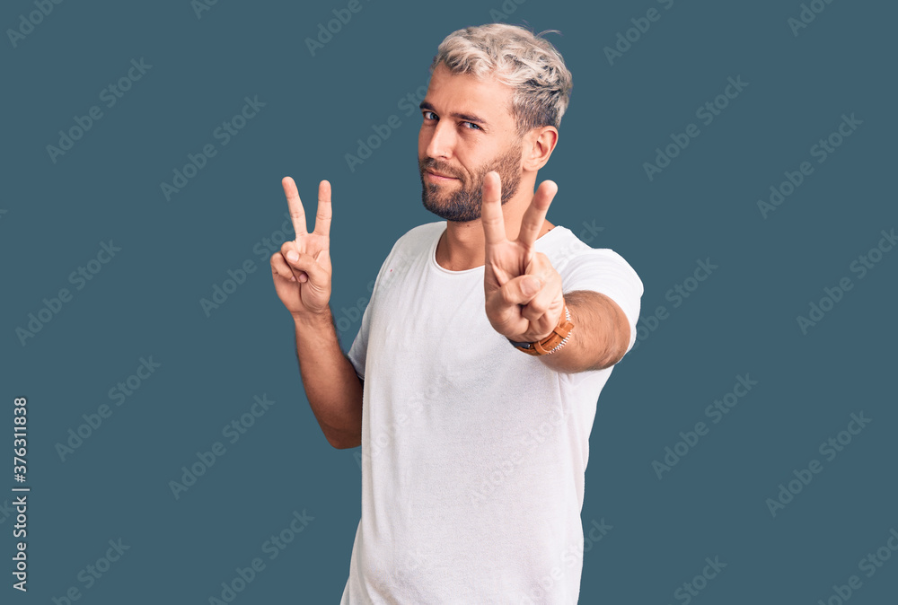 Young handsome blond man wearing casual t-shirt smiling looking to the camera showing fingers doing victory sign. number two. Young handsome blond man wearing casual t-shirt smiling looking to the camera showing fingers doing victory sign. number two.