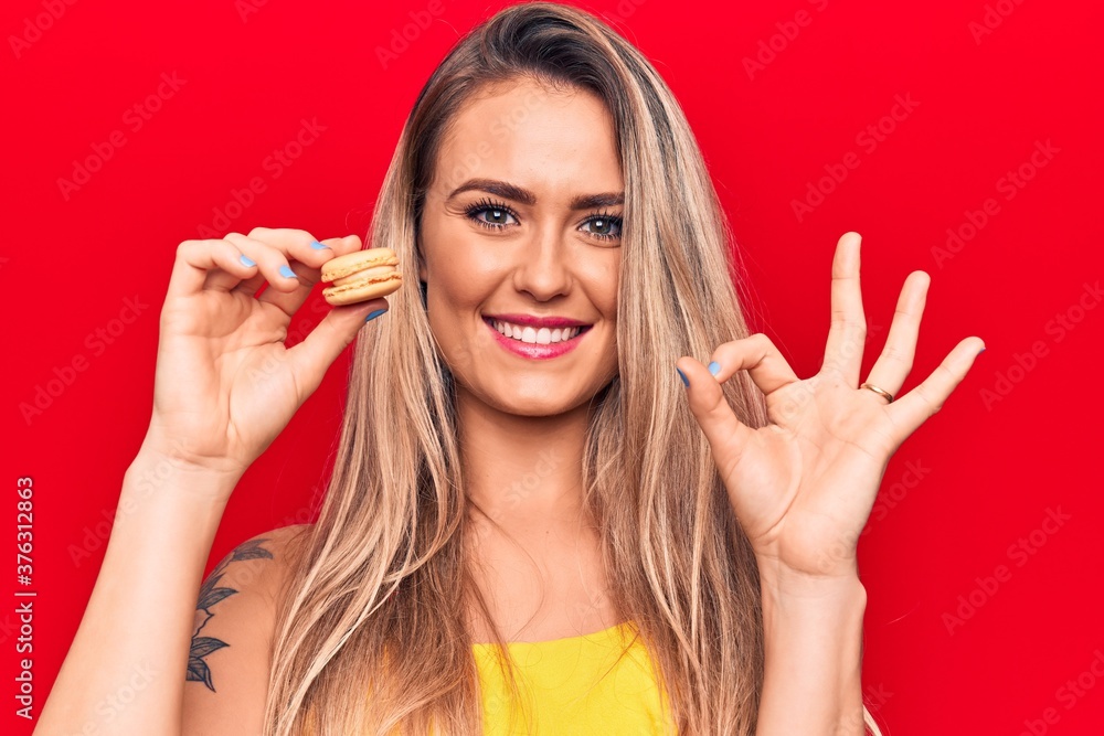 Obraz premium Young beautiful blonde woman eating french dessert macaron over isolated red background doing ok sign with fingers, smiling friendly gesturing excellent symbol