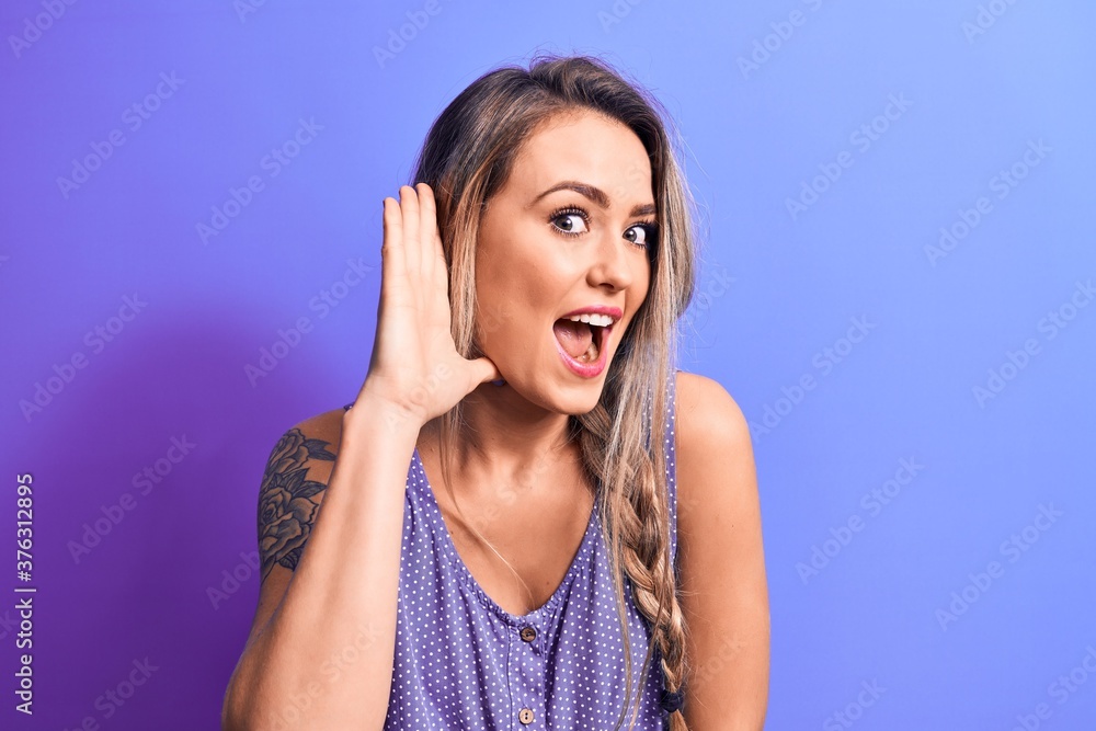 Young beautiful blonde woman wearing casual t-shirt standing over isolated purple background smiling with hand over ear listening and hearing to rumor or gossip. Deafness concept. Young beautiful blonde woman wearing casual t-shirt standing over isolated purple background smiling with hand over ear listening and hearing to rumor or gossip. Deafness concept.