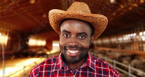 Portrait of handsome cheerful man in hat and motley shirt standing in stable of sheep farm rising face and smiling to camera. Happy male farmer with smile in shed with cattle. Close up.