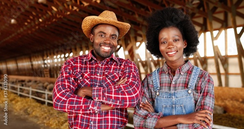 Portrait of happy African American young couple of farmers standing together and crossing hands while smiling to camera in sheep farm shed. Woman and man in stable. Male and female joyful workers.