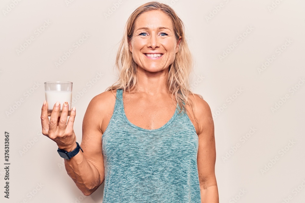 Middle age caucasian blonde woman holding a glass of milk looking positive and happy standing and smiling with a confident smile showing teeth Middle age caucasian blonde woman holding a glass of milk looking positive and happy standing and smiling with a confident smile showing teeth