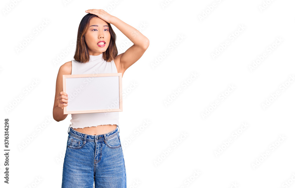 Young beautiful chinese girl holding empty white chalkboard stressed and frustrated with hand on head, surprised and angry face Young beautiful chinese girl holding empty white chalkboard stressed and frustrated with hand on head, surprised and angry face