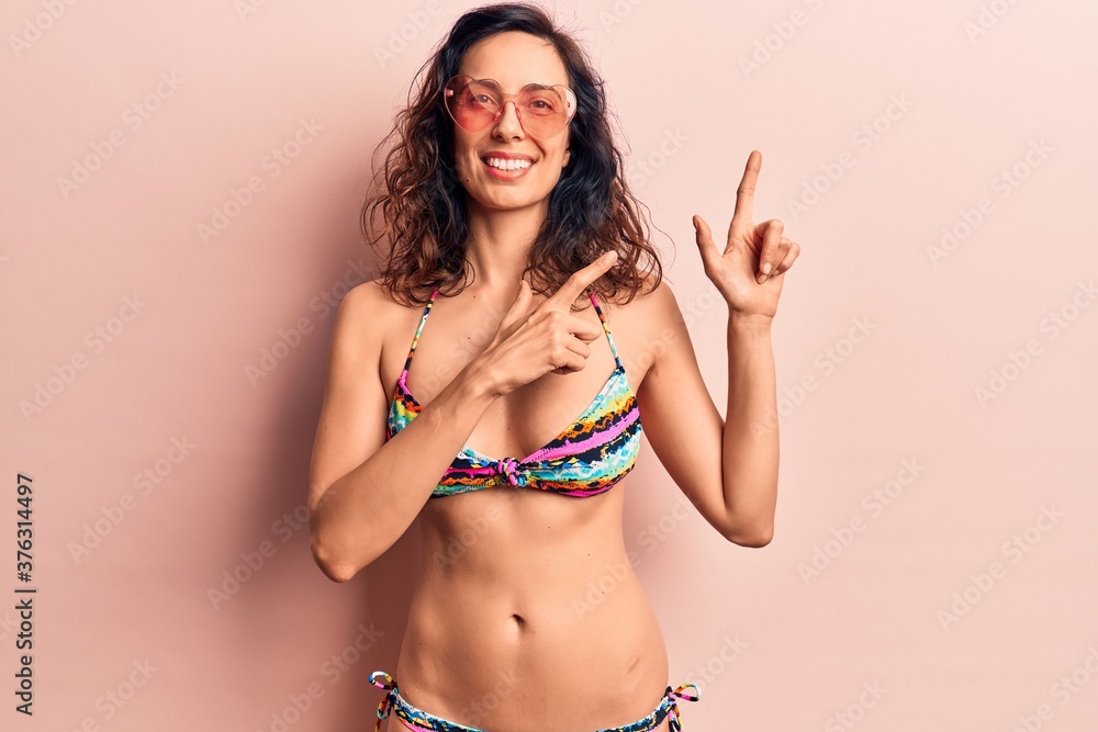 Naklejka premium Young beautiful hispanic woman wearing bikini and heart sunglasses smiling and looking at the camera pointing with two hands and fingers to the side.