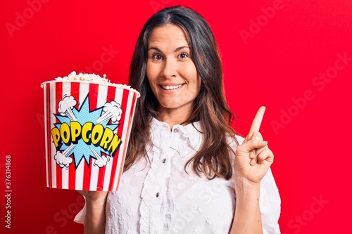 Young beautiful brunette woman eating pack of popcorns over isolated red background smiling happy pointing with hand and finger to the side
