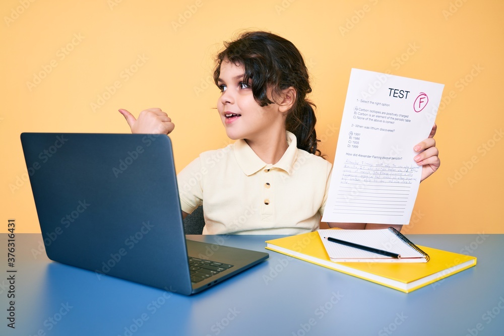Cute hispanic child showing failed exam sitting on the desk pointing thumb up to the side smiling happy with open mouth Cute hispanic child showing failed exam sitting on the desk pointing thumb up to the side smiling happy with open mouth