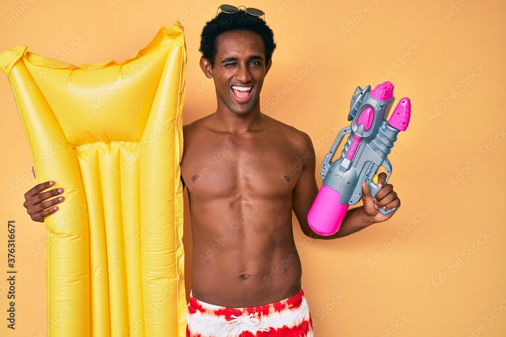 African handsome man wearing swimsuit and holding summer float winking looking at the camera with sexy expression, cheerful and happy face. African handsome man wearing swimsuit and holding summer float winking looking at the camera with sexy expression, cheerful and happy face.