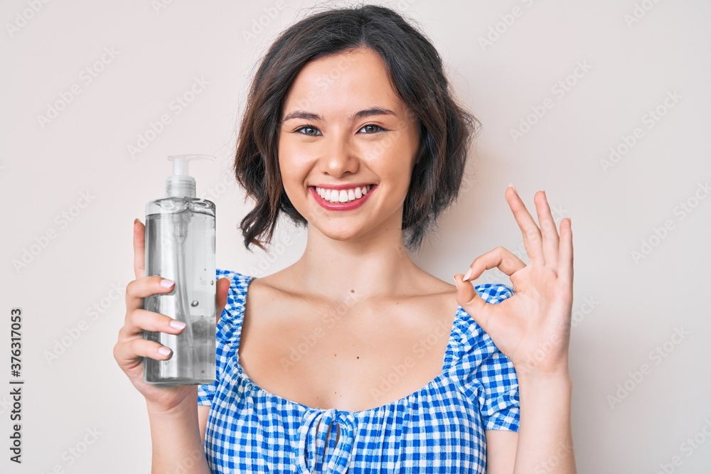 Young beautiful woman holding hand sanitizer gel doing ok sign with fingers, smiling friendly gesturing excellent symbol Young beautiful woman holding hand sanitizer gel doing ok sign with fingers, smiling friendly gesturing excellent symbol