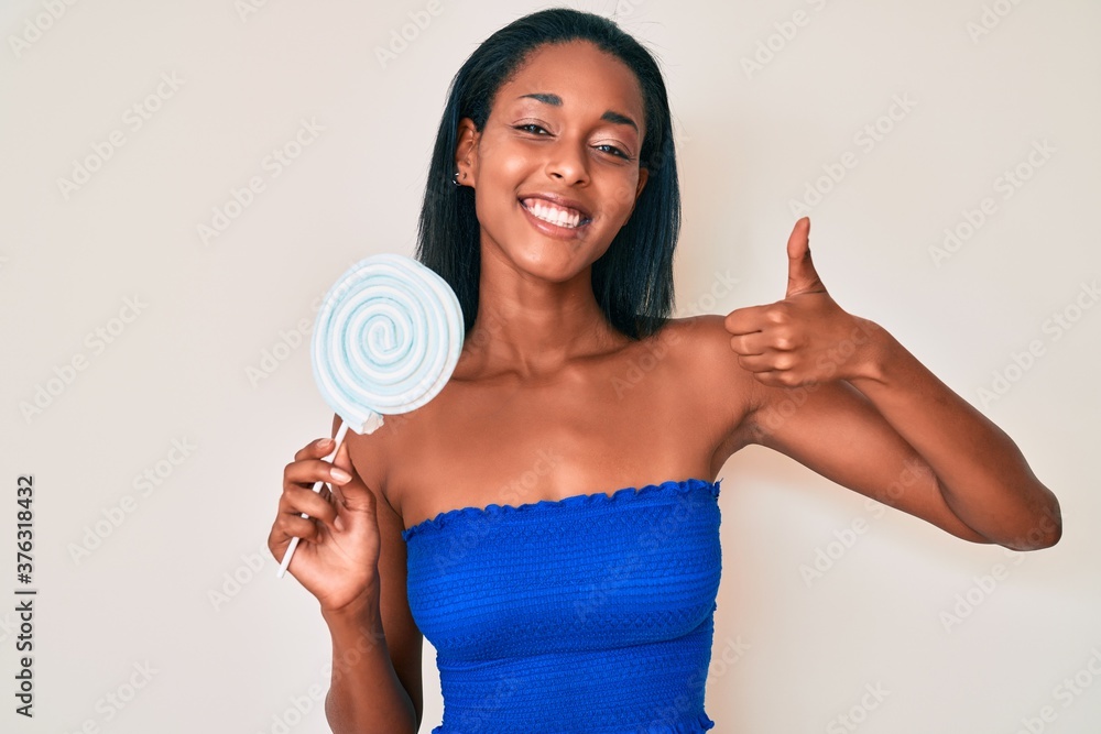 Young african american woman holding sugar candy smiling happy and positive, thumb up doing excellent and approval sign Young african american woman holding sugar candy smiling happy and positive, thumb up doing excellent and approval sign