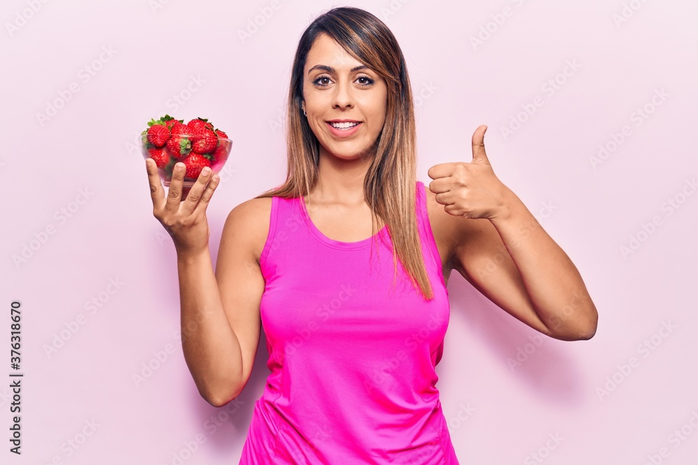 Young beautiful woman holding bowl with strawberries smiling happy and positive, thumb up doing excellent and approval sign Young beautiful woman holding bowl with strawberries smiling happy and positive, thumb up doing excellent and approval sign