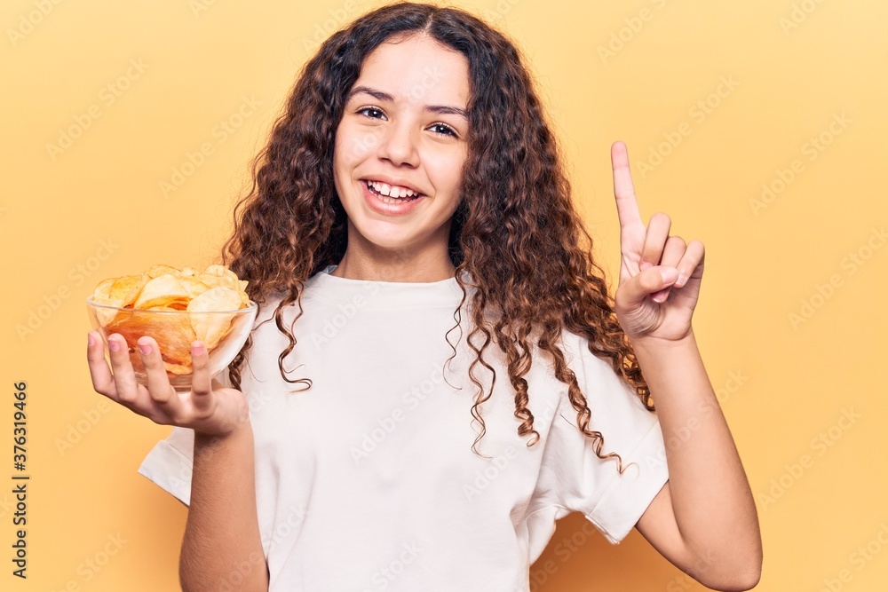 Beautiful kid girl with curly hair holding potato chip smiling with an idea or question pointing finger with happy face, number one Beautiful kid girl with curly hair holding potato chip smiling with an idea or question pointing finger with happy face, number one