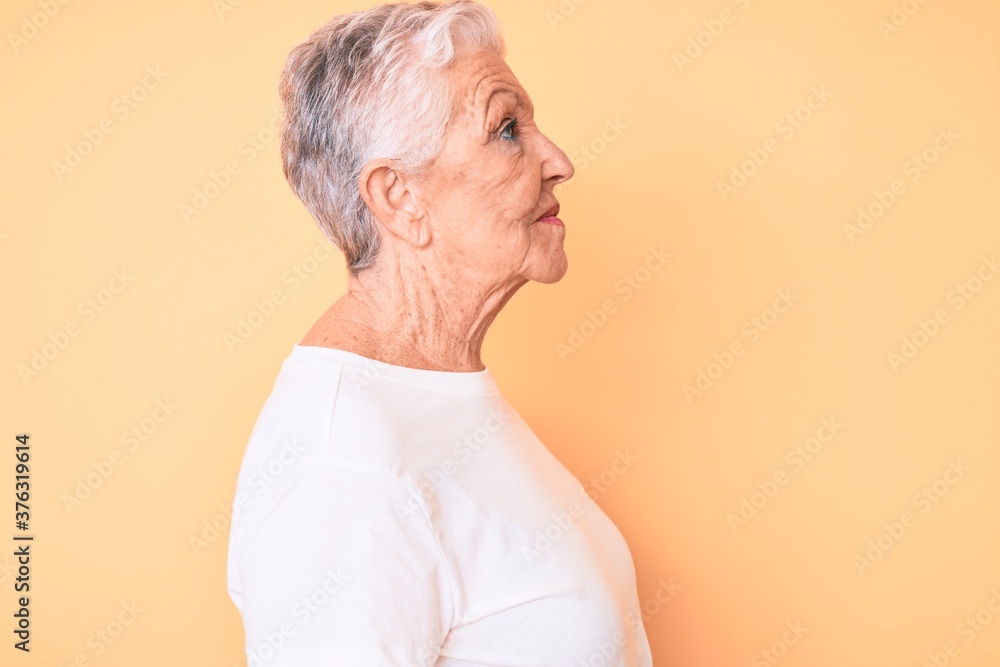 Senior beautiful woman with blue eyes and grey hair wearing classic white tshirt over yellow background looking to side, relax profile pose with natural face with confident smile. Senior beautiful woman with blue eyes and grey hair wearing classic white tshirt over yellow background looking to side, relax profile pose with natural face with confident smile.