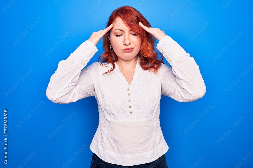 Fototapeta premium Young beautiful redhead businesswoman wearing elegant shirt standing over blue background with hand on head, headache because stress. Suffering migraine.