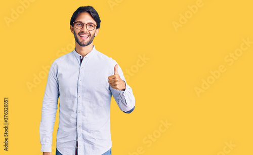 Handsome hispanic man wearing business shirt and glasses doing happy thumbs up gesture with hand. approving expression looking at the camera showing success.