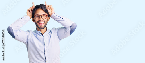 Handsome hispanic man wearing business shirt and glasses doing funny gesture with finger over head as bull horns