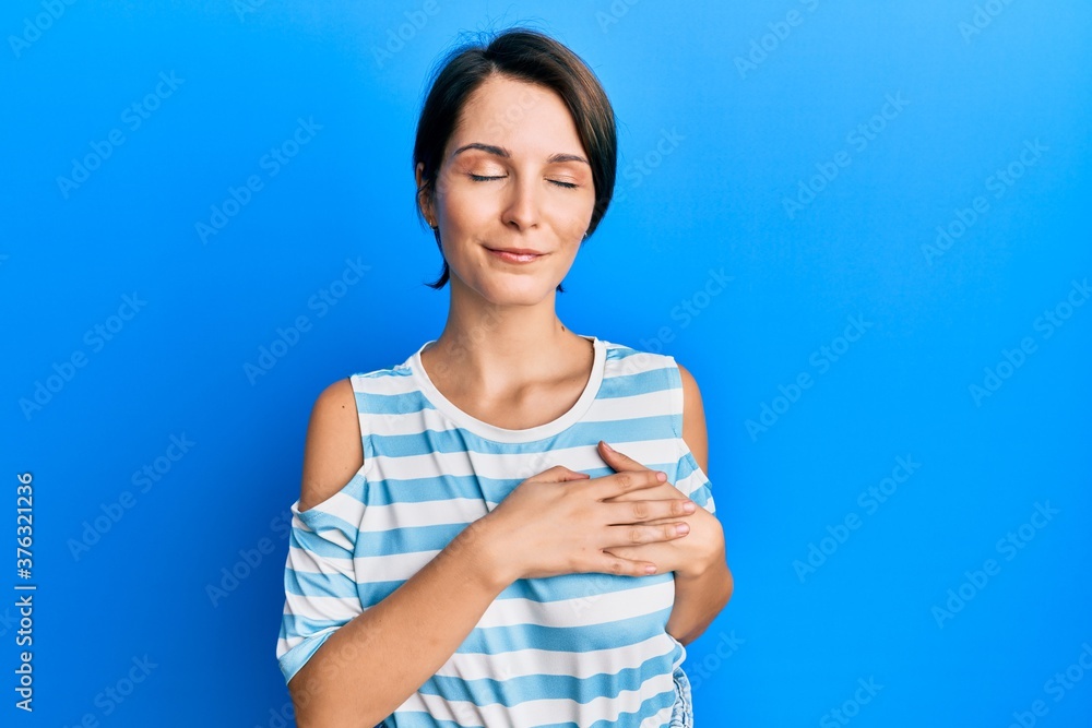Young brunette woman with short hair wearing casual striped t-shirt smiling with hands on chest with closed eyes and grateful gesture on face. health concept. Young brunette woman with short hair wearing casual striped t-shirt smiling with hands on chest with closed eyes and grateful gesture on face. health concept.