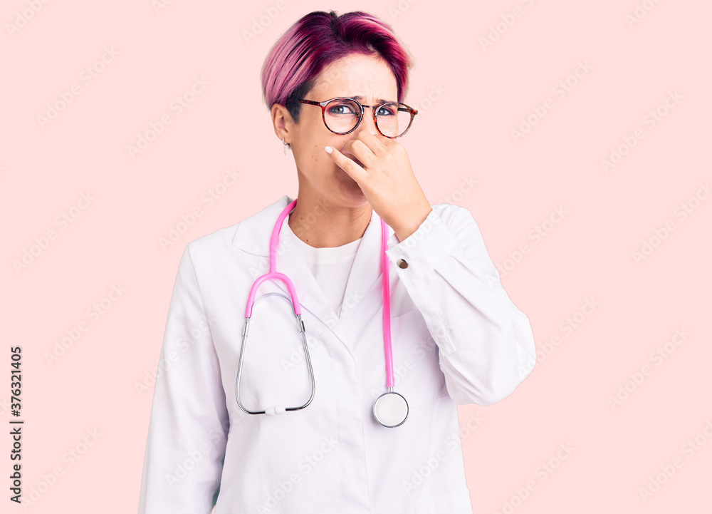 Young beautiful woman with pink hair wearing doctor uniform smelling something stinky and disgusting, intolerable smell, holding breath with fingers on nose. bad smell Young beautiful woman with pink hair wearing doctor uniform smelling something stinky and disgusting, intolerable smell, holding breath with fingers on nose. bad smell