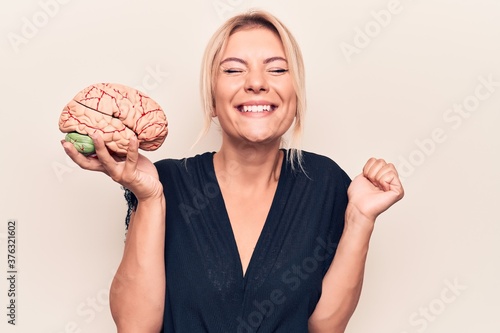 Young beautiful blonde woman asking for care memory holding brain over white background screaming proud, celebrating victory and success very excited with raised arm