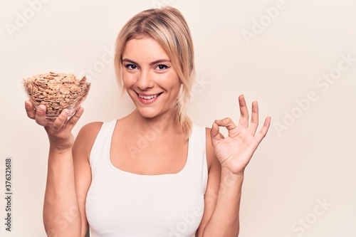 Young beautiful blonde woman holding bowl with conrflakes cerals over white background doing ok sign with fingers, smiling friendly gesturing excellent symbol