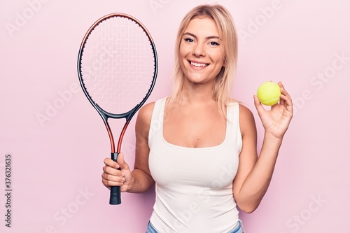 Young beautiful blonde sportswoman playing tennis using racket and ball over pink background looking positive and happy standing and smiling with a confident smile showing teeth © Krakenimages.com