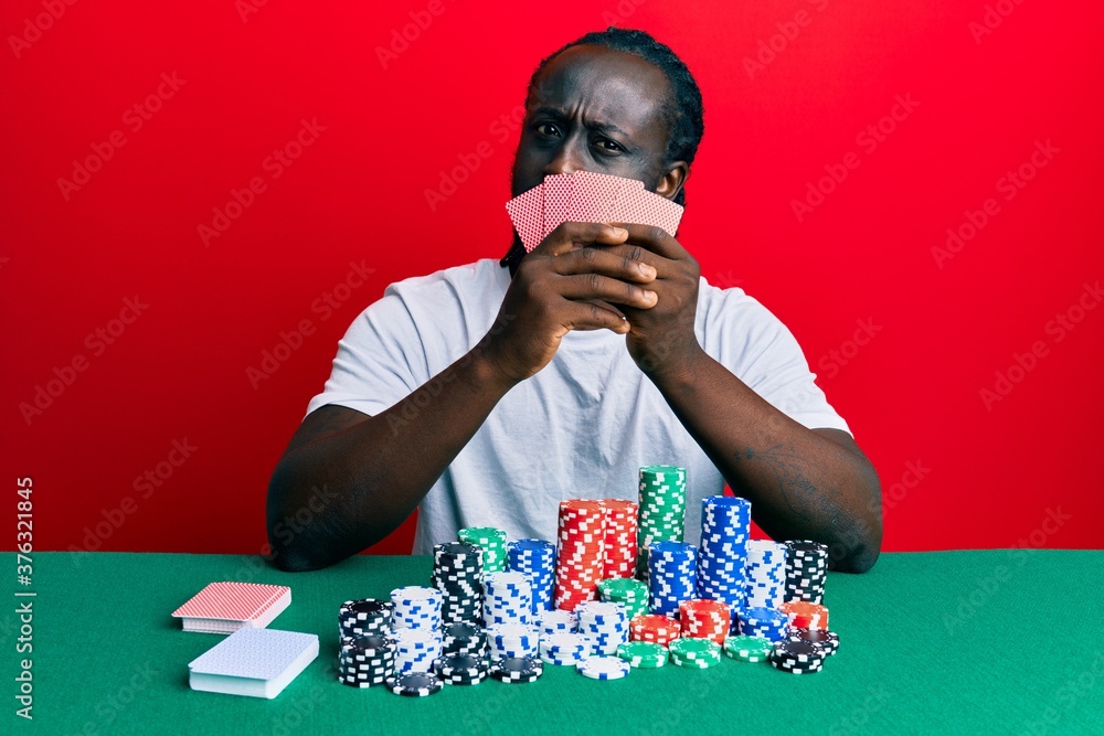 Handsome young black man playing gambling poker covering face with cards in shock face, looking skeptical and sarcastic, surprised with open mouth Handsome young black man playing gambling poker covering face with cards in shock face, looking skeptical and sarcastic, surprised with open mouth