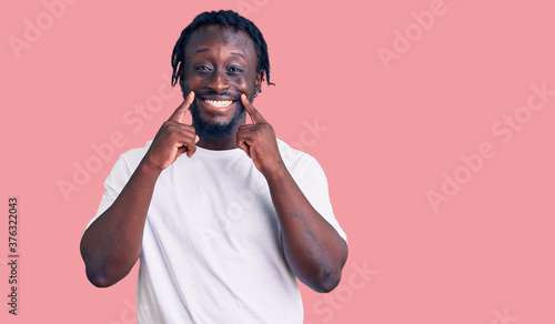 Young african american man with braids wearing casual white tshirt smiling with open mouth, fingers pointing and forcing cheerful smile
