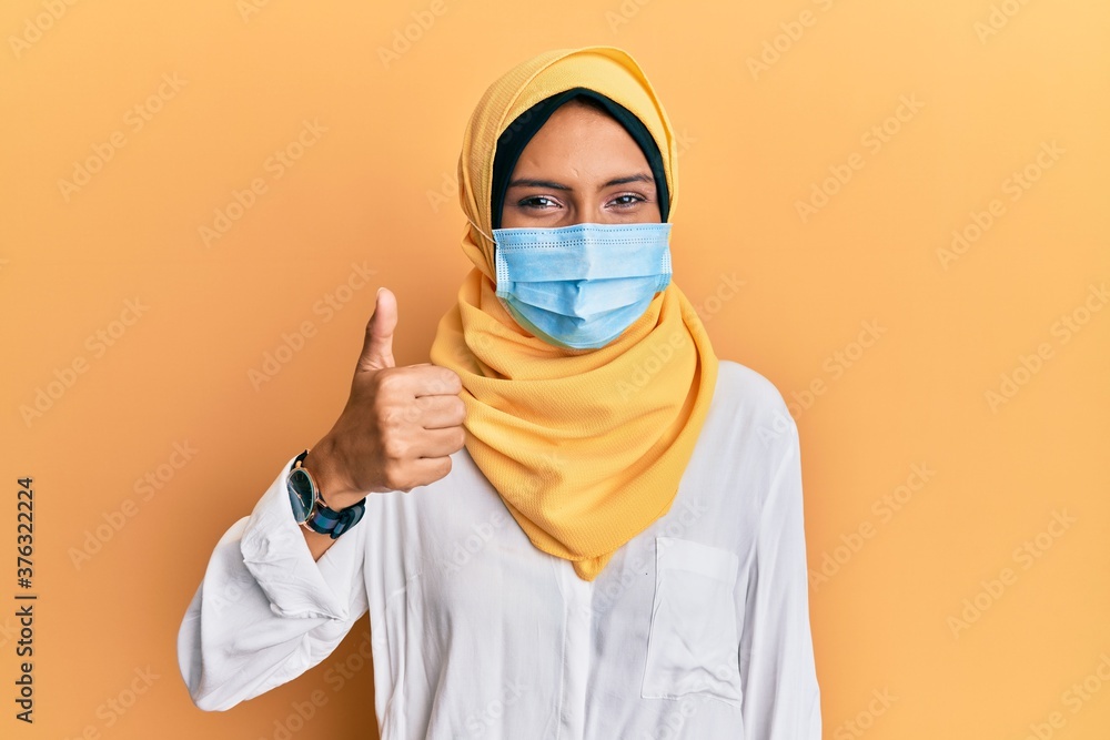 Young brunette arab woman wearing traditional islamic hijab and medical mask smiling happy and positive, thumb up doing excellent and approval sign Young brunette arab woman wearing traditional islamic hijab and medical mask smiling happy and positive, thumb up doing excellent and approval sign