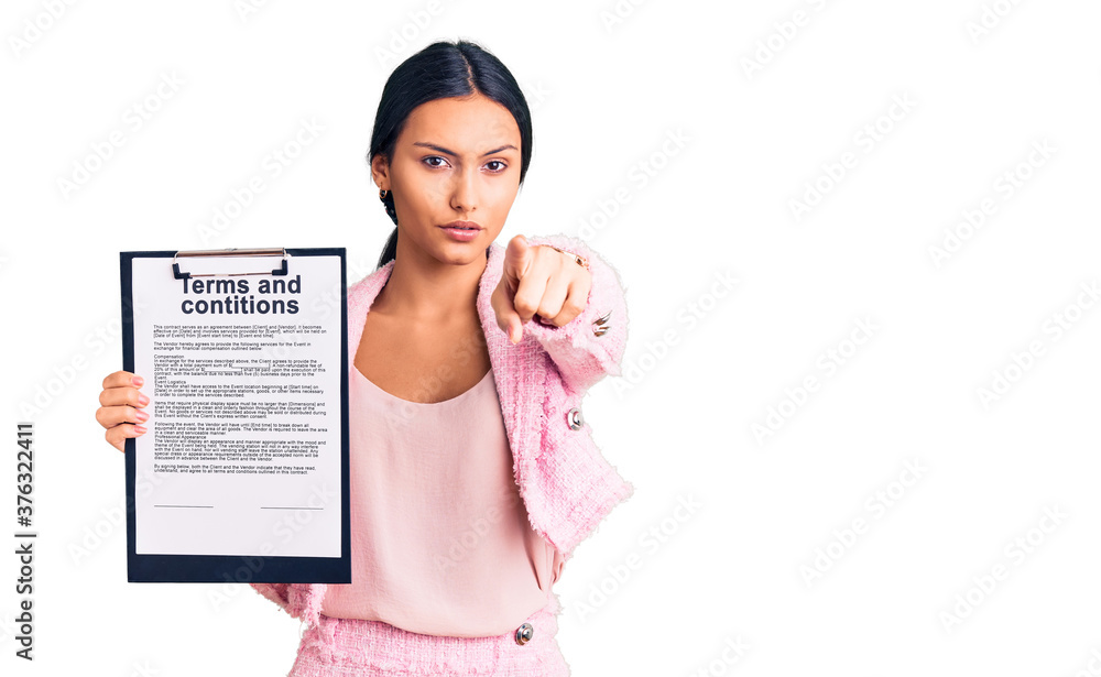 Young beautiful latin girl holding clipboard with terms and conditions document pointing with finger to the camera and to you, confident gesture looking serious Young beautiful latin girl holding clipboard with terms and conditions document pointing with finger to the camera and to you, confident gesture looking serious