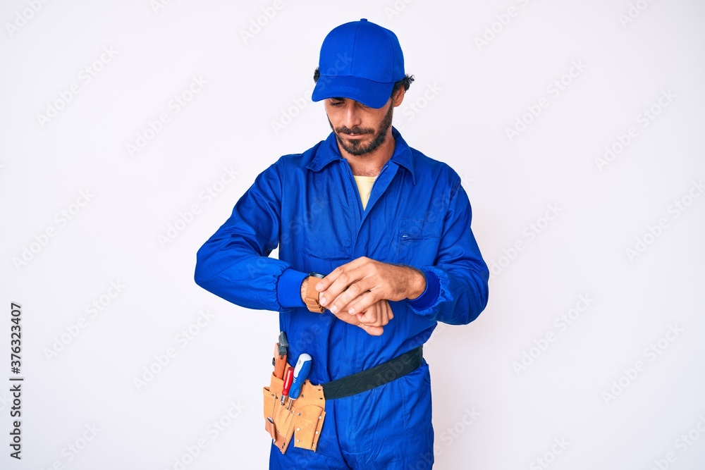 Handsome young man with curly hair and bear weaing handyman uniform checking the time on wrist watch, relaxed and confident