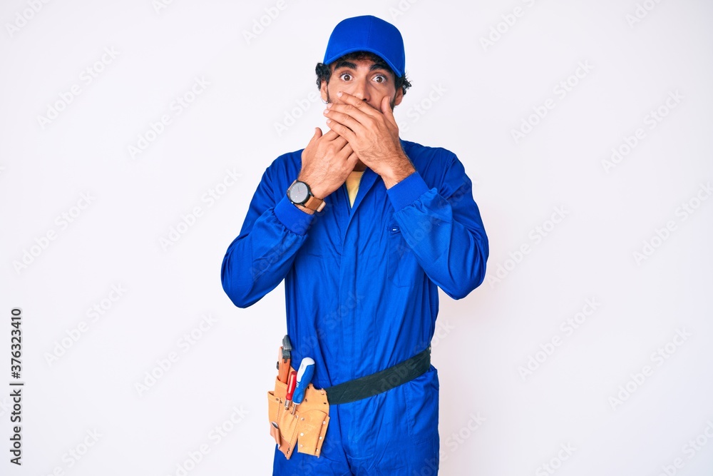 Handsome young man with curly hair and bear weaing handyman uniform shocked covering mouth with hands for mistake. secret concept.