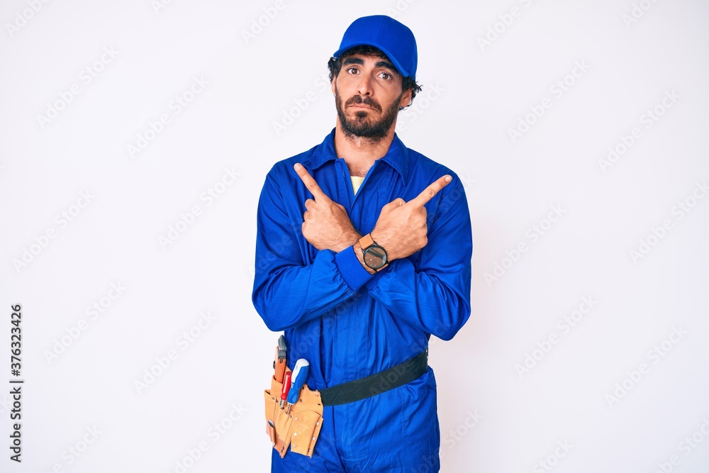 Handsome young man with curly hair and bear weaing handyman uniform pointing to both sides with fingers, different direction disagree