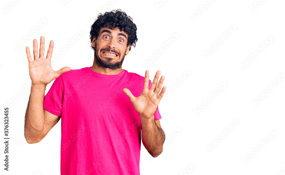 Handsome young man with curly hair and bear wearing casual pink tshirt afraid and terrified with fear expression stop gesture with hands, shouting in shock. panic concept. Handsome young man with curly hair and bear wearing casual pink tshirt afraid and terrified with fear expression stop gesture with hands, shouting in shock. panic concept.