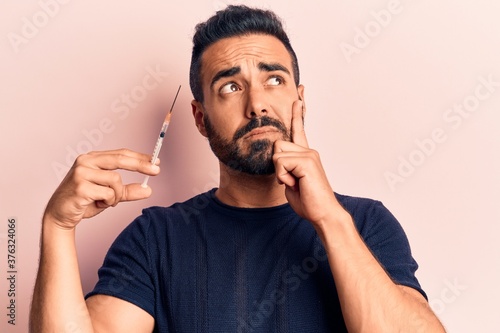 Young hispanic man holding syringe serious face thinking about question with hand on chin, thoughtful about confusing idea