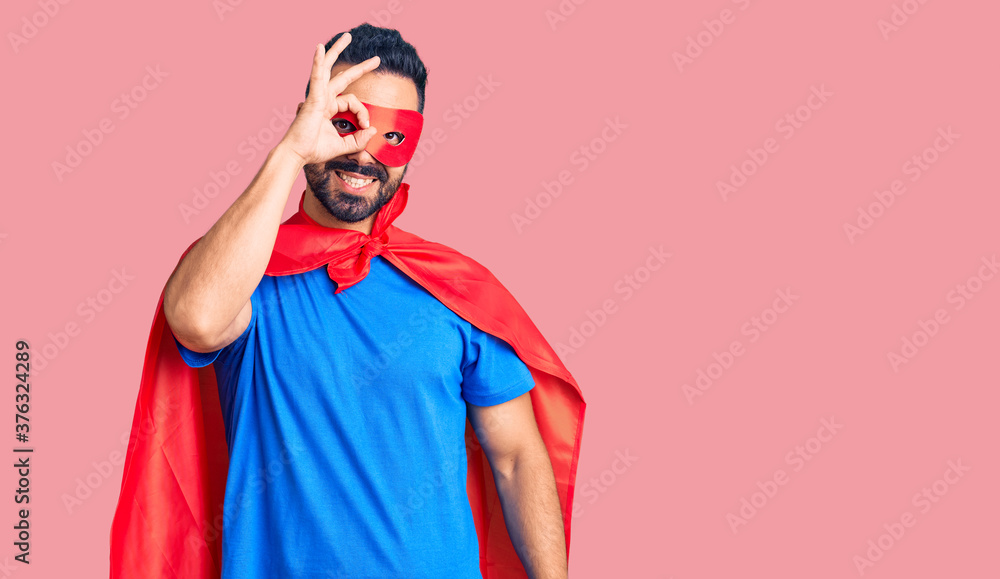 Young hispanic man wearing super hero costume smiling happy doing ok sign with hand on eye looking through fingers Young hispanic man wearing super hero costume smiling happy doing ok sign with hand on eye looking through fingers