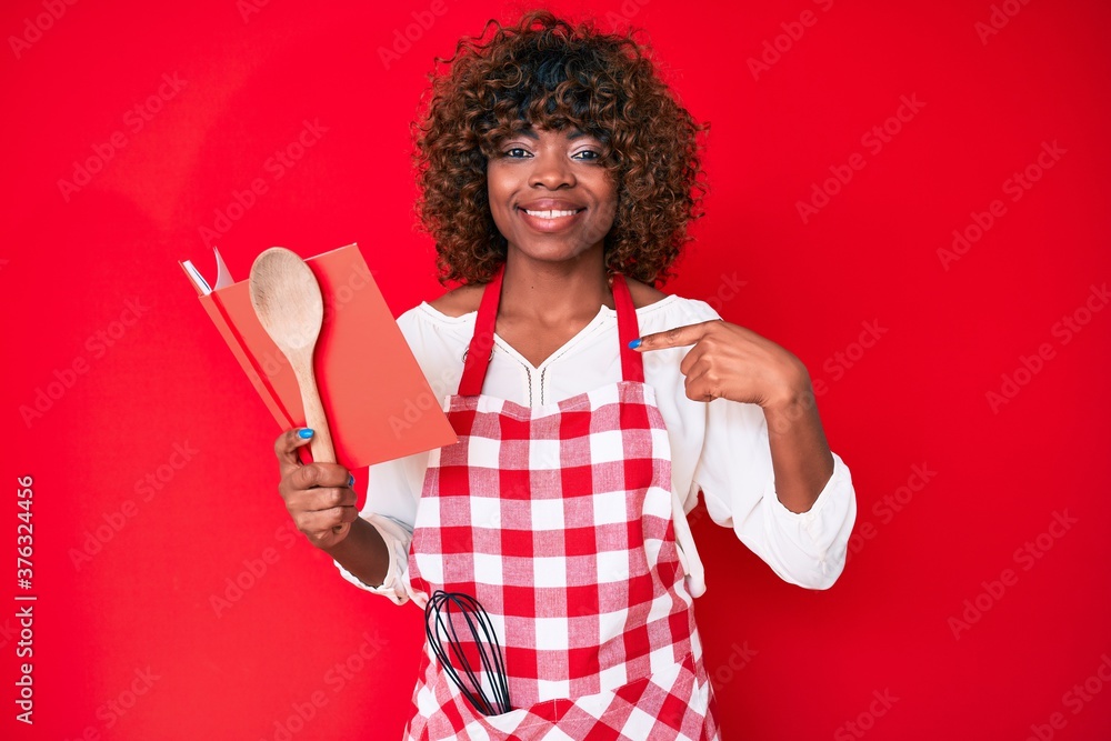 Young african american woman wearing professional baker apron reading cooking recipe book pointing finger to one self smiling happy and proud Young african american woman wearing professional baker apron reading cooking recipe book pointing finger to one self smiling happy and proud