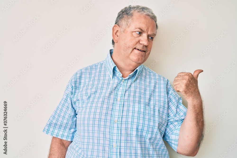 Senior grey-haired man wearing casual clothes smiling with happy face looking and pointing to the side with thumb up. Senior grey-haired man wearing casual clothes smiling with happy face looking and pointing to the side with thumb up.