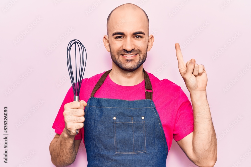 Young handsome bald man wearing apron holding whisk smiling with an idea or question pointing finger with happy face, number one Young handsome bald man wearing apron holding whisk smiling with an idea or question pointing finger with happy face, number one