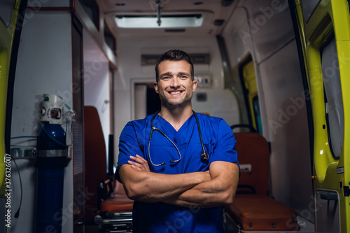 A portrait of a young intern standing in front of an ambulance car with his hands folded and smiling. © Anna Kosolapova