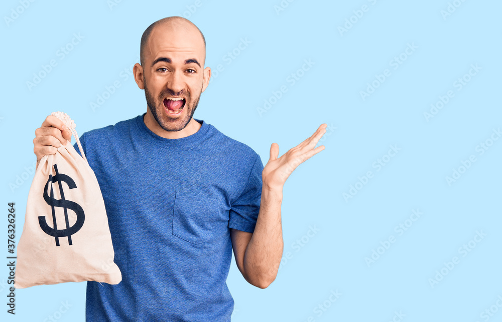 Young handsome man holding money bag with dollar symbol celebrating victory with happy smile and winner expression with raised hands Young handsome man holding money bag with dollar symbol celebrating victory with happy smile and winner expression with raised hands