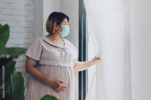 Pregnant Asian women wear a protective mask to prevent germs. Look out of the window During the quarantine at home for preventing coronavirus and covid-19 infection.