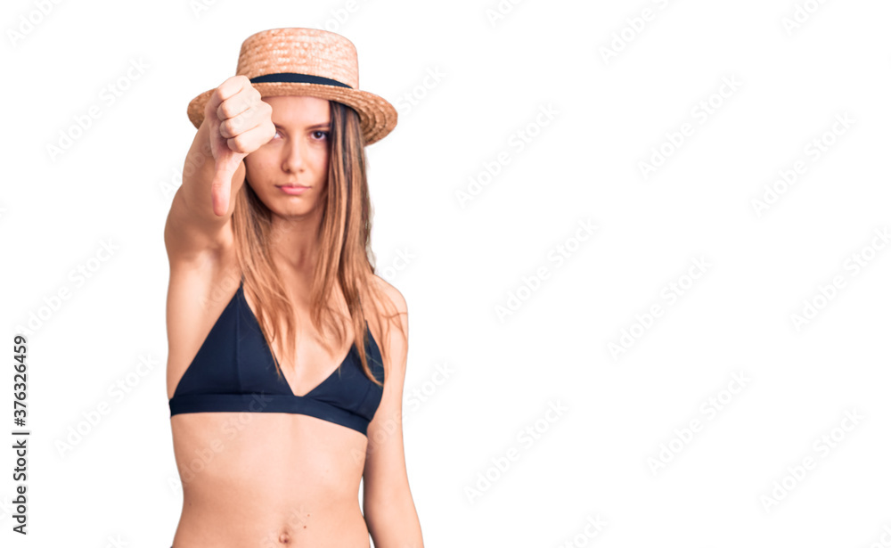 Young beautiful girl wearing bikini and hat looking unhappy and angry showing rejection and negative with thumbs down gesture. bad expression. Young beautiful girl wearing bikini and hat looking unhappy and angry showing rejection and negative with thumbs down gesture. bad expression.