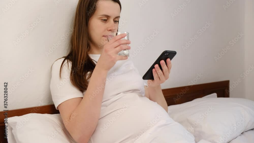 Pregnant woman enjoying using mobile apps texting typing messages sit on comfortable white bed, drinking fresh water, future mom holding smartphone looking at cellphone screen browsing social media Pregnant woman enjoying using mobile apps texting typing messages sit on comfortable white bed, drinking fresh water, future mom holding smartphone looking at cellphone screen browsing social media