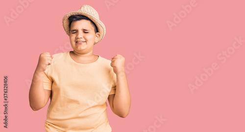 Little boy kid wearing summer hat and hawaiian swimsuit excited for success with arms raised and eyes closed celebrating victory smiling. winner concept.