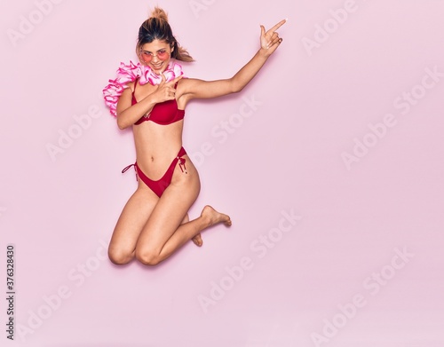 Young beautiful girl on vacation wearing bikini and hawaiian lei smiling happy. Jumping with smile on face pointing with fingers to the side over isolated pink background