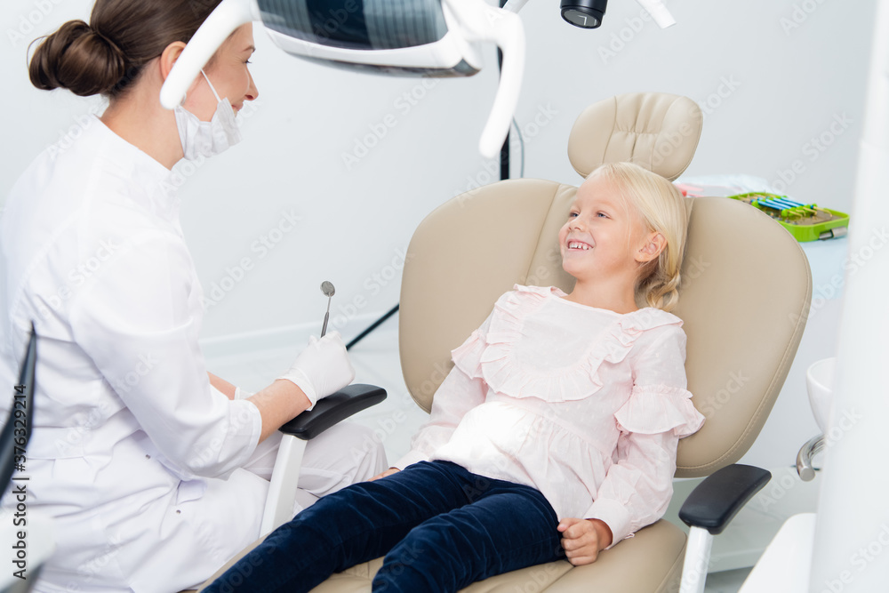 A satisfied little dentist patient showing her perfect smile after dental treatment in a clinic. A box with medical tools in the background. A satisfied little dentist patient showing her perfect smile after dental treatment in a clinic. A box with medical tools in the background.