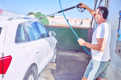 Middle age handsome man wearing casual clothes and sunglasses smiling happy. Standing with smile on face washing car using water pistol. © Krakenimages.com